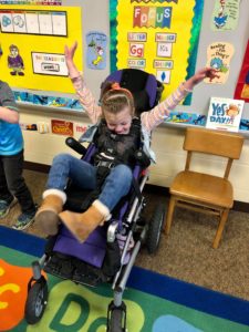 Sadie in her adaptive stroller in her classroom with arms in the air and a smile on her face.