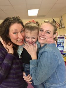 Sadie smiling with her teachers holding her up in between them in her classroom.