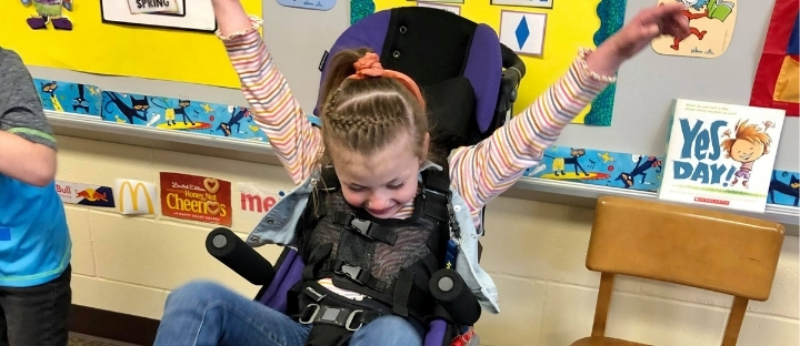 Sadie in her adaptive stroller in her classroom with her arms in their air and a smile on her face